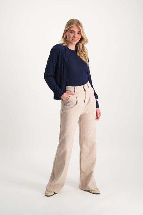 Het model staat in een witte studio en draagt de Loiza trousers in de kleur Cream. De Loiza trousers is een broek met hoge taille en wijd uitlopende pijpen. De broek is van een zachte stof gemaakt.